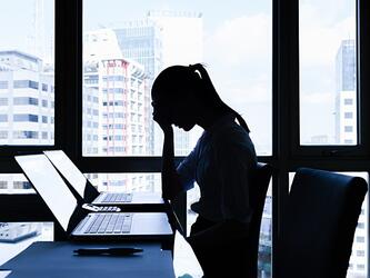 stressed woman at a desk with head bowed over laptop, with tall buildings in background